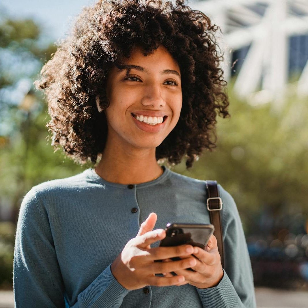 A woman with curly hair smiling while using her smartphone outdoors.